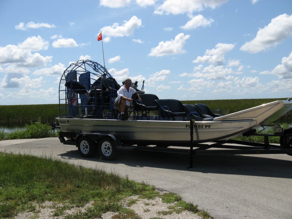 An airboat parked on a trailer at the edge of the Everglades, with a guide sitting on board under a sunny sky.