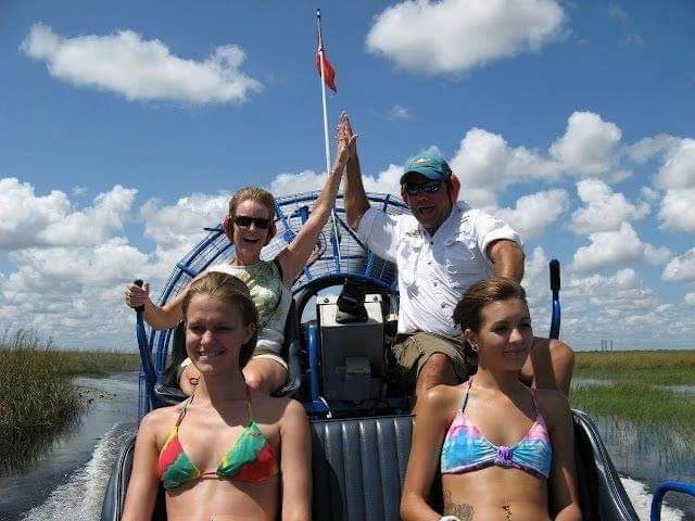 A group of happy tourists enjoying an exciting airboat ride through the Everglades, with the captain and a passenger high-fiving under a bright blue sky